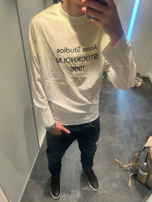Acne studios tröja  - Vit långärmad topp från Acne Studios med svart texttryck 'STOCKHOLM 1996' på bröstet. Klassisk rund halsringning och relaxed fit. Perfekt för dig som gillar minimalistisk och stilren design.