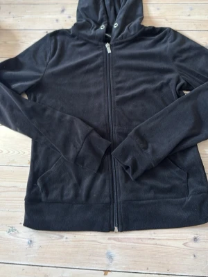 Svart zip hoodie Gina  - En klassisk svart hoodie med dragkedja framtill och två fickor. Hoodien har huva med snörning och långa ärmar. Perfekt för en avslappnad och stilren look. Materialet känns mjukt och bekvämt.