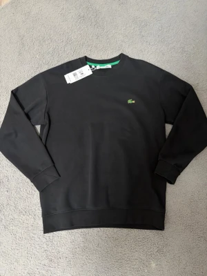 Svart sweatshirt från Lacoste - Klassisk svart sweatshirt från Lacoste med rund halsringning och det ikoniska gröna krokodilmärket broderat på bröstet. Tröjan har ribbade muddar vid ärmslut och nederkant samt en enkel, stilren design. Perfekt för en avslappnad och trendig look.