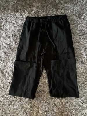 Svarta Essentials mjukisbyxor - Säljer ett par svarta mjukisbyxor från Essentials Fear of God. Byxorna har dragsko i midjan, raka ben och diskret logga på vänster lår. Tillverkade i mjukt och bekvämt material, perfekta för chill dagar eller streetwear-stil.