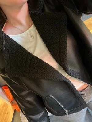 Svart teddyjacka med dragkedja - Säljer en svart teddyjacka med bred krage och detaljer i mjukt teddyfoder. Jackan har en oversized passform, synliga sömmar och silverfärgad dragkedja framtill samt på fickorna. Perfekt för dig som vill ha en trendig och mysig jacka till hösten eller vintern. 