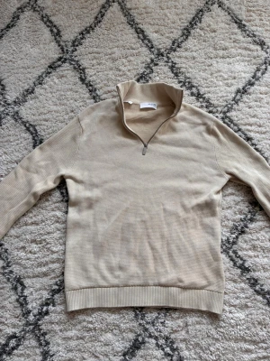 Beige halvzip tröja med ribbade detaljer - Stilren beige tröja med halvzip och hög krage. Tröjan har diskret struktur i materialet och ribbade muddar vid ärmslut och nederkant. Perfekt för lager på lager och enkel att matcha med olika outfits.