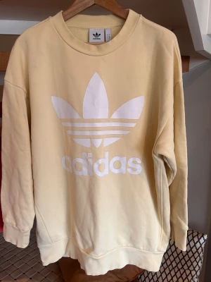 Gul oversized sweatshirt från Adidas - Säljer en ljusgul sweatshirt från Adidas med stor vit Trefoil-logga och text på bröstet. Tröjan har rund halsringning, långa ärmar och ribbade muddar. Tillverkad i mjukt bomullsmaterial som är skönt mot huden. Perfekt för en avslappnad streetwear-look.