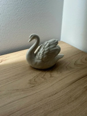 Vit porslinssvan prydnad - Säljer en elegant vit prydnad i form av en svan, tillverkad i porslin. Perfekt som dekoration på hyllan eller bordet. Svanen har fina detaljer på vingarna och en mjuk, glansig yta.