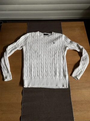 Vit kabelstickad tröja från polo Ralph lauren - Snygg vit kabelstickad tröja med rund hals och långa ärmar. Tröjan har ribbade muddar vid ärmslut och nederkant samt ett diskret broderat emblem på bröstet. Perfekt för lager på lager och passar till det mesta. Materialet ser ut att vara mjukt och bekvämt.