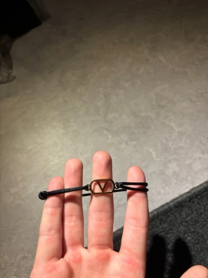 Valentino armband - Snyggt svart armband med justerbart band och en guldfärgad metallbricka i mitten formad som ett V. Perfekt accessoar för dig som gillar minimalistisk och stilren design. Passar till många olika outfits och är lätt att bära.