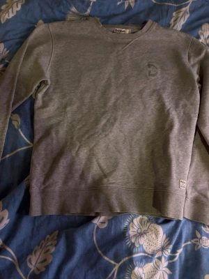Grå sweatshirt från Dobber - Klassisk grå sweatshirt från Dobber med rund halsringning och diskret logga på bröstet. Tröjan har ribbade muddar vid ärmslut och nederkant samt en liten patch nertill. Perfekt för en avslappnad och stilren look. (Är öppen för bud)