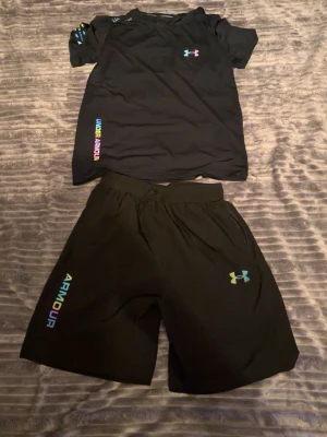 Svart träningsset från Under Armour - Säljer ett svart träningsset från Under Armour med t-shirt och shorts. Båda plaggen har färgglada loggor och detaljer i regnbågsfärger. Materialet är lätt och andas, perfekt för träning eller sportaktiviteter. Elastisk midja på shortsen för skön passform.