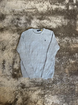 🩵Ljusgrå kabelstickad tröja Polo Ralph Lauren (Large) - ✨Snygg ljusgrå kabelstickad tröja från Polo Ralph Lauren med rund halsringning och klassisk marinblå logga broderad på bröstet. Tröjan har ribbade muddar vid ärmslut och nederkant och är tillverkad i mjukt material som passar perfekt till höst och vinter. Storlek: Large✨