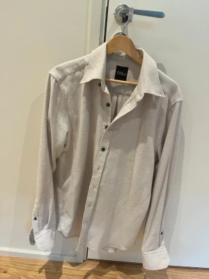 Beige skjorta från Oscar Jacobson - Säljer en stilren beige skjorta från Oscar Jacobson i regular fit. Skjortan har klassisk krage, långa ärmar och knappar framtill. Materialet känns mjukt och har en lätt struktur, perfekt för en clean och avslappnad look.