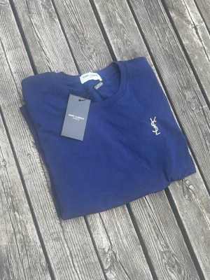 Marinblå Saint Laurent t-shirt - Snygg marinblå t-shirt från Saint Laurent med klassisk rund halsringning och ikonisk YSL-logga broderad i vitt på bröstet. Tillverkad i mjuk bomull som känns skön mot huden. Perfekt för dig som gillar stilrena och exklusiva plagg.
