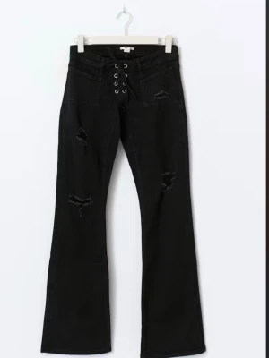Svarta bootcut jeans med snörning - Säljer ett par svarta bootcut jeans med snörning och metallöljetter framtill. Jeansen har slitna detaljer på benen och stora fickor fram. Materialet är klassisk denim med stretch. Snygg passform som markerar midjan och ger en retrovibe.