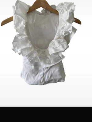 Volangtopp - Snygg vit topp med djup urringning och stora volangdetaljer runt halsen. Ärmlös modell i mjukt material, perfekt för att skapa en trendig och romantisk look. Passar till både jeans och kjol.