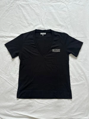 Ganni t-shirt - Svart v-ringad topp från Ganni med broderad logga på bröstet. Har en liten slits i nederkanten på sidan. Begagnat skick, har ett litet hål på baksidan 