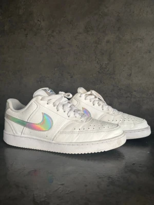 Nike sneakers - Säljer ett par vita Nike sneakers med en snygg holografisk swoosh på sidan. Skorna har klassisk låg profil, perforerad tåbox och vita snören. Ovandelen är i läder och sulan är vit med Nike-logga undertill. Perfekta för dig som vill sticka ut lite extra.