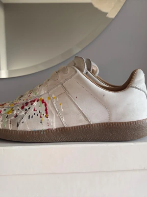 Vita maison margiela med färgstänk - Säljer dessa maison margielas eftersom dem har blivit för små.  Storlek 40 men passar 41.  Pris kan diskuteras. Unika vita sneakers med färgstänk i flera färger som rött, gult, blått och grönt. Skorna har en klassisk låg siluett, vita snören och beige sula. Materialet ser ut att vara mocka och läder, vilket ger en schysst kontrast till de kreativa färgdetaljerna. 