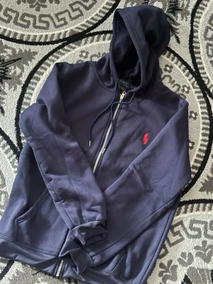 Hoodie Polo Ralph Lauren Slim Fit Zip-Up  - Klassisk marinblå huvtröja med dragkedja och smal passform från Polo Ralph Lauren, prydd med den ikoniska, röda broderade logotypen föreställande en polospelare på bröstet. Tillverkad i ett bekvämt fleecematerial med mjuk insida – perfekt för lager-på-lager-klädsel.  Skick: 10/10 Färg: Marinblå  Snabb och omsorgsfull frakt