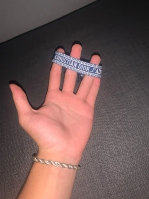 Blått Christian Dior J'Adior armband - Säljer ett blått tygarmband från Christian Dior med texten 'J'ADIOR' broderat i vitt. Armbandet har justerbar knytning med två stora tofsar i ändarna. Perfekt accessoar för att lyfta din outfit med en lyxig touch.