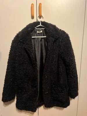 Svart teddyjacka med krage - Mysig svart teddyjacka med fluffig struktur och klassisk krage. Jackan har två knappar framtill och är helfodrad med slät insida. Perfekt för dig som vill ha en varm och trendig look under kyliga dagar.