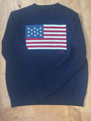 Ralph Lauren tröja med amerikanska flaggan - Marinblå Ralph Lauren-tröja med amerikanska flaggan på framsidan. Klassisk modell med rund hals, långärmad och ribbade muddar. Storlek M. Begagnad och i fint skick. Se bilder för skick och detaljer.