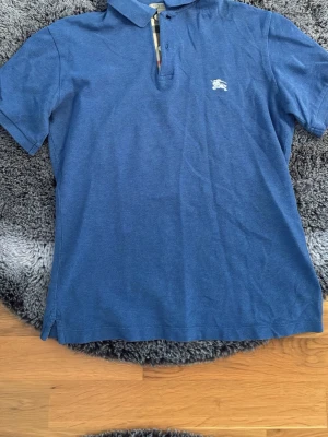 Blå pikétröja med knappslå - Snygg blå pikétröja i bomull med klassisk krage och knappslå framtill. Tröjan har korta ärmar och en liten broderad logga på bröstet. Perfekt för en avslappnad och stilren look.