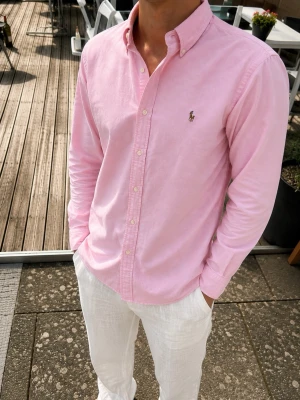 Ralph Lauren Oxford Skjorta – Rosa Slim Fit M - Snygg och klassisk Ralph Lauren Oxford-skjorta i ljusrosa. 👔 • Slim Fit • Storlek M • 100% bomull • Klassisk button-down krage • Broderad Polo-logo på bröstet • Stilren rosa färg • Mycket fint skick  Perfekt skjorta till både vardag, sommaren och lite finare outfits. Snygg att bära tillsammans med exempelvis vita linnebyxor, chinos eller jeans.