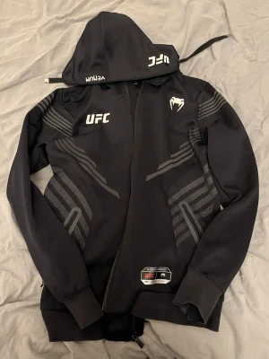 Svart UFC hoodie med dragkedja - Säljer en svart hoodie från Venum med UFC-logga och snygga gråa detaljer på ärmar och sidor. Tröjan har huva med snörning, dragkedja framtill och muddar vid ärmslut. Perfekt för träning eller chill. Materialet är mjukt och stretchigt.