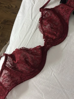 Vinröd spets-bh med bygel - Säljer en vinröd bh i spets med bygel och tunna axelband. Bh:n har vackra blommiga spetsdetaljer och ger en feminin look. Perfekt för dig som gillar eleganta underkläder med fina detaljer. Storlek 70B men skulle säga att den är lite mindre än så. Aldrig använd då den var alldeles för liten.