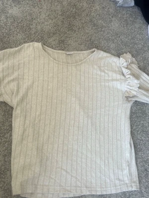 Beige ribbad topp med volangärm - Säljer en beige ribbad topp med rund hals och korta ärmar. Båda ärmerna har en snygg volangdetalj som ger toppen en unik touch. Perfekt att styla med jeans eller kjol för en trendig look. Stor i storleken!