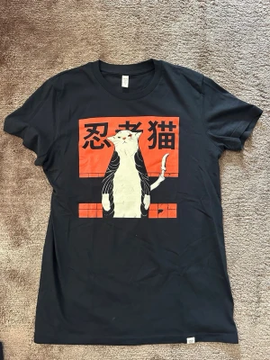 Svart t-shirt med ninja-katt från Wituka - Cool svart t-shirt från Wituka i ekologisk bomull. Framsidan har ett stort tryck med en ninja-katt och japanska tecken på röd bakgrund. Klassisk rund hals och korta ärmar. Perfekt för dig som gillar unika och lekfulla motiv.