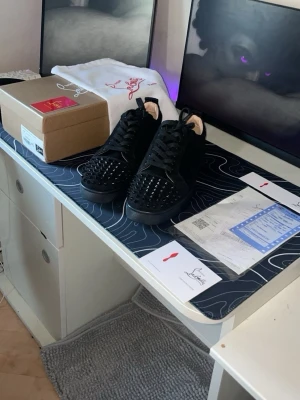 Svarta sneakers med nitar - Säljer ett par svarta sneakers från Christian Louboutin med svarta nitar på tån och ovansidan. Skorna har svart mocka, svarta skosnören och en klassisk låg siluett. Perfekta för dig som vill sticka ut med en edgy och exklusiv look.