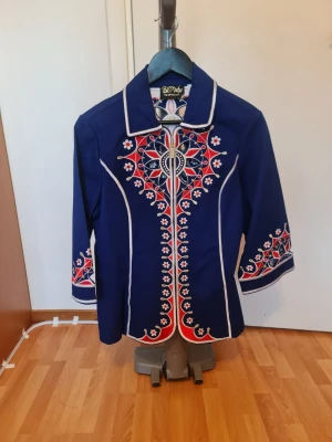 Blå skjorta med broderi och krage - Unik blå skjorta med vit och röd broderad design framtill, baktill och på ärmarna. Skjortan har markerad krage, vita kantdetaljer och trekvartsärmar. Perfekt för dig som vill sticka ut med färg och mönster.