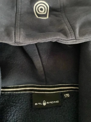 Mörkblå hoodie från Sail Racing - Mörkblå hoodie från Sail Racing med dragkedja och broderad logga på bröstet. Hoodien har en bröstficka med dragkedja och mjukt fleecefoder på insidan. Klassisk huva och sportig design, perfekt för dig som gillar stilrena detaljer. Köpte den för 1000 mitt pris är 180 jättebra pris för skicket som den är i jag vill bara ha bort den och käna lite snabba pengar.