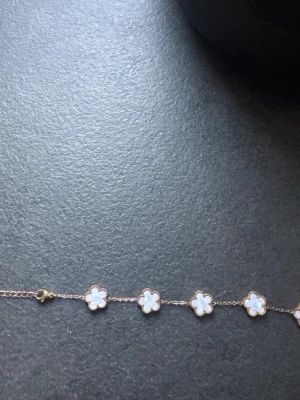 Guldfärgat armband med vita blommor - Snyggt armband i guldfärg med små vita blommor i pärlemor-look. Blommorna sitter tätt längs hela kedjan och ger en söt och elegant känsla. Perfekt accessoar för att lyfta vilken outfit som helst.