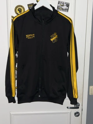Svart AIK tröja - Säljer en svart träningsjacka med gula ränder längs ärmarna och AIK-emblem på bröstet. Jackan har dragkedja framtill, ståkrage och två fickor med dragkedja. Från mentalita. 50kr går till aiktifo.