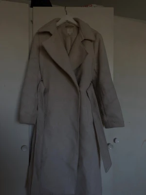 Beige lång kappa med skärp - Stilren beige kappa i lång modell med bred krage och tillhörande skärp i midjan. Kappan har diskreta fickor på sidorna och en klassisk, rak passform. Perfekt för dig som vill ha en enkel men snygg look till hösten eller vintern.