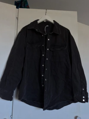 Svart overshirt med fickor - Stilren svart overshirt med klassisk krage och tryckknappar framtill. Två stora bröstfickor med lock och knapp, samt knappdetaljer vid ärmslut. Overshirten har en avslappnad passform och är tillverkad i ett mjukt bomullsliknande material.
