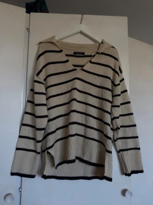 Beige stickad tröja med svarta ränder - Snygg beige stickad tröja med svarta horisontella ränder och v-ringning. Tröjan har avslappnad passform, långa ärmar och ribbade muddar. Perfekt för lager på lager och chill stil. Tillverkad i mjukt stickat material.