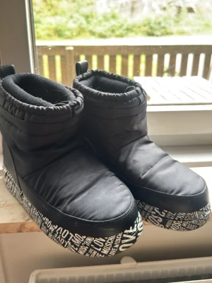 Svarta Moon Boots från Love Moschino - Säljer ett par svarta Moon Boots från Love Moschino med cool textmönstrad sula och stort rött hjärta med logga bak. Boots i vadderat material och dragflik baktill. Perfekta för vinterns alla äventyr!