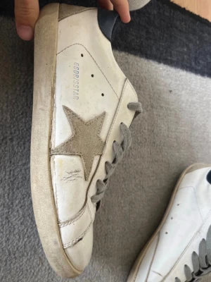 Golden goose superstar  - Säljer ett par vita superstars från Golden Goose med grå mockastjärna på sidan, grå skosnören och mörkblå detalj vid hälen. Skorna har en klassisk låg profil och är tillverkade i läder med mockadetaljer. Perfekta för en avslappnad look.