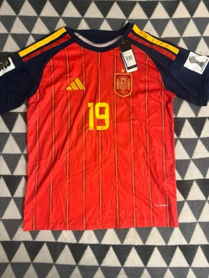 Spansk landslagströja Adidas röd M - Säljer en röd matchtröja från Spaniens landslag, Adidas, med gula detaljer och nummer 19 på bröstet. Tröjan har blå ärmar med gula och röda ränder på axlarna samt broderad landslagssköld. Tillverkad i lätt funktionsmaterial, slim fit.