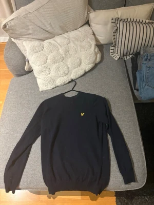 Mörkblå stickad tröja med gul logga - Snygg mörkblå stickad tröja med rund hals och lång ärm. Tröjan har en liten gul broderad logga på bröstet och ribbade muddar vid ärmslut och nederkant. Perfekt att matcha med jeans eller kjol.