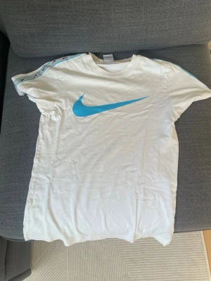 Vit Nike t-shirt med blå logga - Säljer en vit t-shirt från Nike med stor blå swoosh-logga på bröstet och blå detaljer längs axlarna. Klassisk rund hals och korta ärmar. Perfekt för dig som gillar sportig och clean stil.