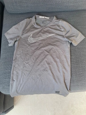 Grå Nike Pro Dri-Fit t-shirt - Säljer en grå Nike Pro Dri-Fit t-shirt med korta ärmar och stor vit Swoosh-logga på bröstet. Tröjan är tillverkad i ett lätt och ventilerande funktionsmaterial som passar perfekt till träning och sport.