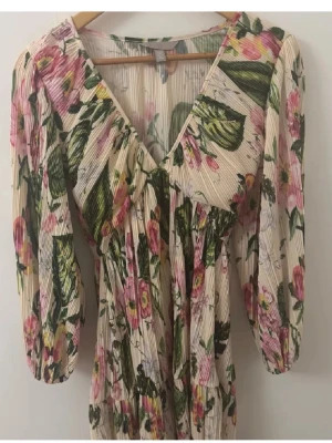 Blommig plisserad kortklänning - Söt kortklänning med plisserat tyg i ljusbeige ton och blommigt mönster i rosa, grönt och gult. Klänningen har v-ringning, trekvartsärm och volangdetaljer. Perfekt för sommarens alla tillfällen.