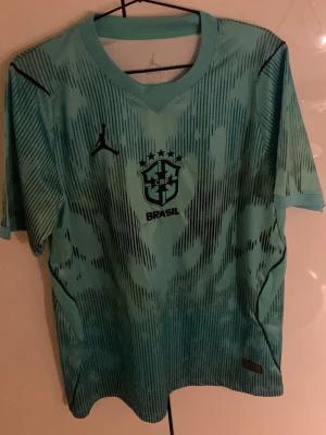 Brasilien tröja  - Säljer en snygg fotbollströja från Brasil med Jordan-logga på bröstet. Tröjan har ett unikt mönster i olika nyanser av neongrönt/blått och svart, rund halsringning och korta ärmar. Perfekt för dig som älskar fotboll och vill sticka ut på planen.