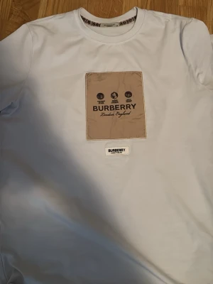 Vit t-shirt från Burberry - Snygg vit t-shirt från Burberry med beige patch och logga på bröstet. Klassisk rund halsringning och korta ärmar. Tillverkad i mjuk bomull som känns skön mot huden. Perfekt för dig som gillar stilrena plagg med exklusiv känsla.
