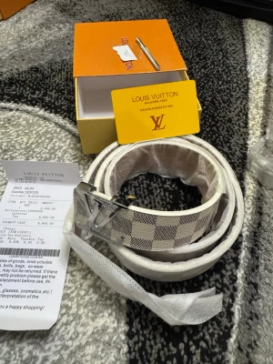Vit rutig LV bälte  - Säljer ett snyggt vitt bälte från LV med klassiskt rutigt mönster och en stor LV-logga i silver på spännet. Skärpet har en exklusiv känsla och passar perfekt till att lyfta vilken outfit som helst.