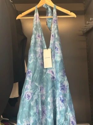 Blommig blå långklänning från Adoore - Säljer en blå långklänning från Adoore med blommigt mönster i lila och blå toner. Klänningen har halterneck och öppen rygg. Oanvänd med prislapp på. 