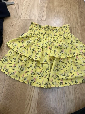 Gul blommig volangkjol från Seaside - Superfin gul kortkjol från Seaside med blommigt mönster i rosa och grönt. Kjolen har bred smock i midjan och dubbla volanger för en extra söt look. Perfekt för varma dagar och ger en riktigt somrig vibe.